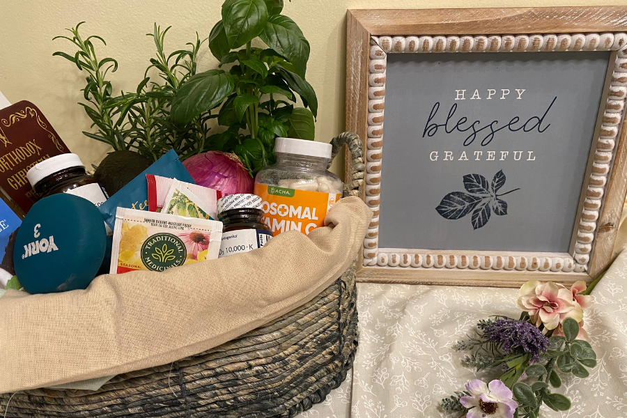 wellness basket filled with items herbs, prayer book, weight, tea, vitamins, onion and avocado next to a sign